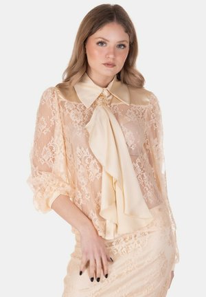 Giovane donna con capelli ondulati che indossa una camicetta di pizzo crema trasparente con grande colletto e fiocco legato al collo, gonna di pizzo abbinata, sfondo neutro.