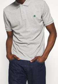 Harmaa, lyhythihainen polo-paita, jossa on kaulus, kahdella napilla varustettu tyylikäs sulkeminen ja vihreä brodeeratun logon. yhdistetty tummansinisiin farkkuihin.