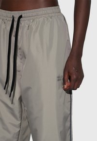 Ljusgrå träningsbyxor med elastisk midja, svart dragsko, sidfickor och ton-i-ton logotypdetalj. Har en slät, något blank yta.