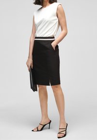 Bílé top bez rukávů, černá tužková sukně s bočním rozparkem a černé sandály na vysokém podpatku se strappy. Drží černou psaníčko. Hladká tkanina.