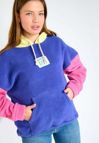 Pullover in fleece blu con accenti rosa e gialli. Presenta una tasca a canguro e un'etichetta decorativa. Texture morbida, vestibilità rilassata.