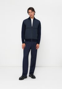 Gilet zippé bleu marine avec des panneaux matelassés à l'avant, des manches ajustées et un col haut, associé à un pantalon bleu marine et des chaussures noires.