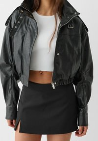 Veste cropped en simili cuir noir avec fermeture éclair, boutons-pression aux épaules et ourlet élastique, associée à un crop top texturé blanc et une mini jupe noire.