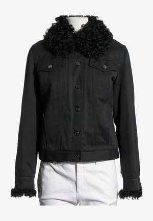 Zwarte jas met knopen, met krullende bontkraag en manchetten, gedragen over witte broek op een paspop tegen een witte achtergrond.