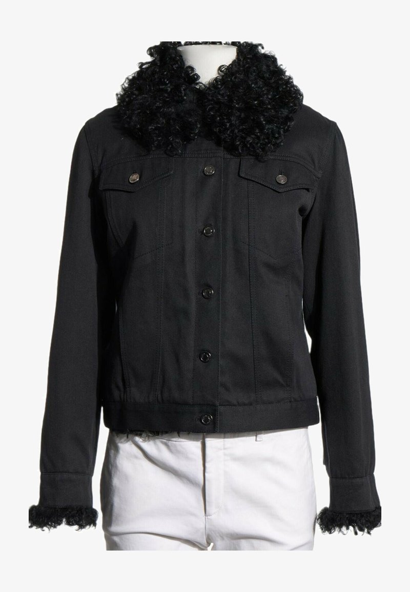 Veste noire boutonnée avec col et poignets en fourrure bouclée, portée sur un pantalon blanc sur un mannequin, sur fond blanc.