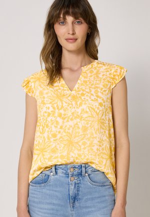 Femme portant un chemisier jaune à fleurs, manches courtes et col en V, rentré dans un jean en denim bleu clair, sur un fond uni.