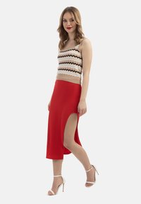 Gonna midi a coste rossa con spacco laterale, abbinata a un top senza maniche a zigzag beige e marrone. Il modello indossa sandali con tacco a strappo bianchi.