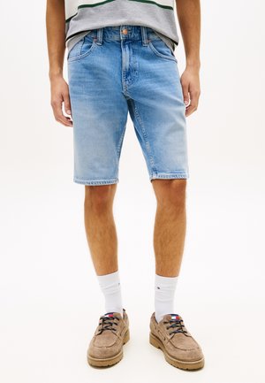 Mand iført lyseblå denimshorts, hvide crew-sokker og beige ruskindssnøresko, stående mod en ensartet lys baggrund.
