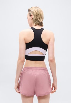 Vrouw met kort blond haar, die een zwart-witte sportbh en roze sportshort draagt, staand met haar rug naar de camera tegen een effen lichte achtergrond.