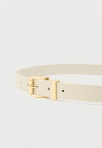 Ζώνη από μπεζ δέρμα με χρυσή μεταλλική πόρπη, που διαθέτει πολλαπλές τρύπες για ρύθμιση μεγέθους και ανάγλυφο λογότυπο στην πόρπη.