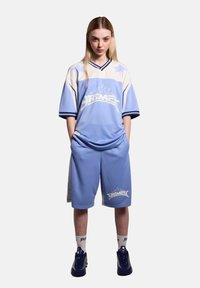 Jersey azul claro y pantalones cortos a juego, hechos de un tejido transpirable, con acentos blancos y un logo gráfico. Se lleva con zapatillas azules.