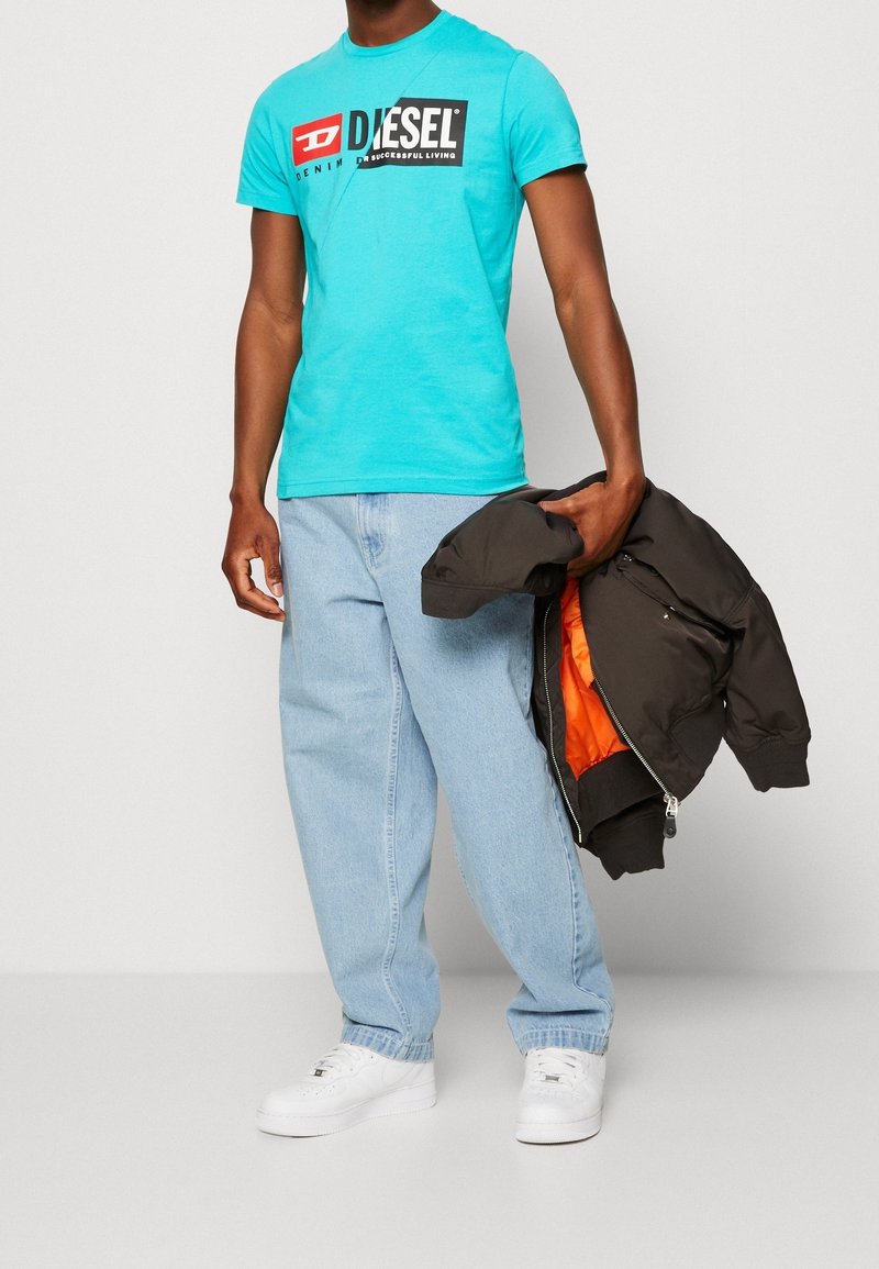 Homme portant un t-shirt turquoise Diesel, un jean bleu clair, des baskets blanches, tenant une veste noire avec une doublure orange dans sa main droite.
