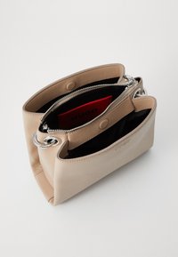 Sac à main en cuir beige au design structuré, avec des accessoires en argent, des fermetures éclair et un intérieur noir contrastant avec un étiquette de marque rouge.
