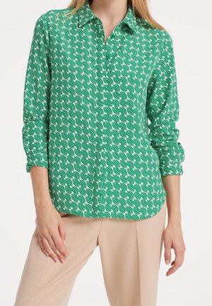 Femme portant une chemise à manches longues verte et blanche à motifs avec un pantalon beige, debout avec une main posée sur sa cuisse.