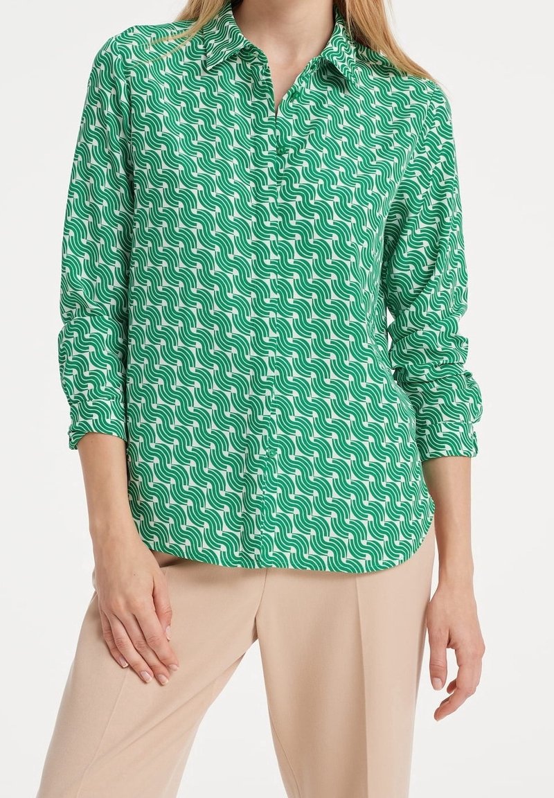 Femme portant une chemise à manches longues verte et blanche à motifs avec un pantalon beige, debout avec une main posée sur sa cuisse.