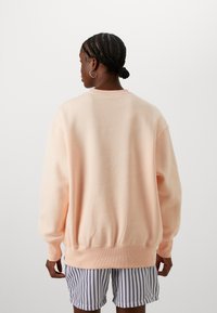 Sudadera melocotón con un corte relajado, puños y dobladillo acanalados. Lleva puesta sobre pantalones cortos a rayas, mostrando una textura suave y un diseño sencillo.