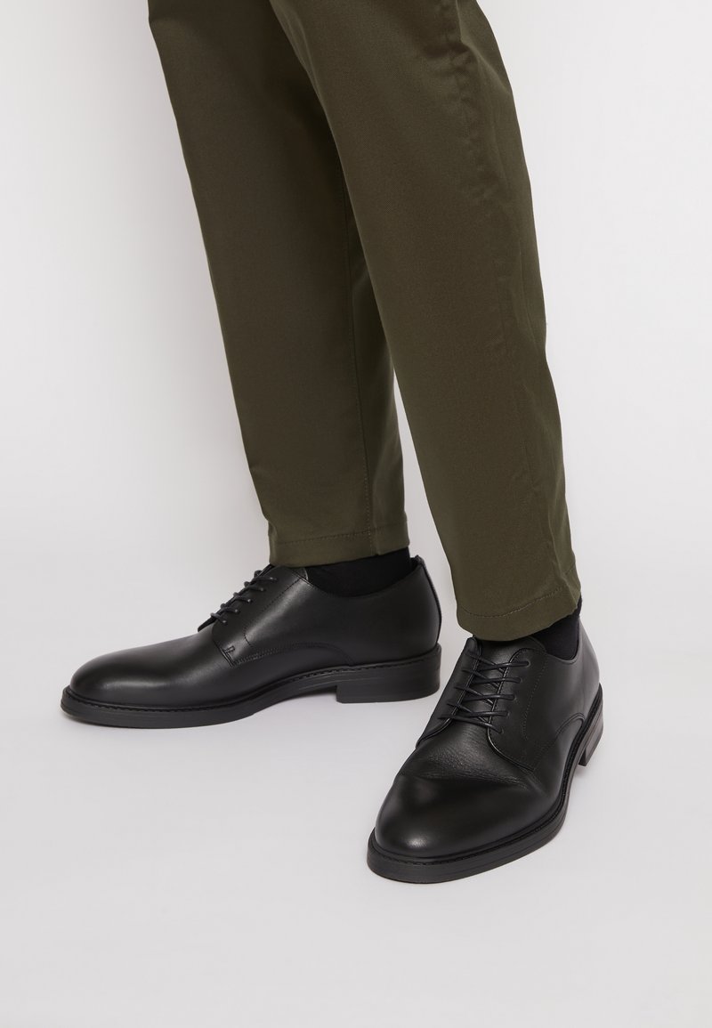 Chaussures habillées en cuir noir avec une finition lisse, un design lacé et un bout arrondi. Associées à un pantalon verde olive.