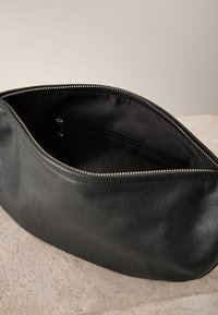 Pochette en cuir noir avec une fermeture éclair métallique partiellement ouverte, montrant un intérieur en tissu noir et une poche intérieure zippée, posée sur une surface beige texturée.
