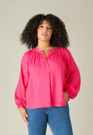 Blouse fuchsia avec un col froncé, des boutons sur le devant et des manches ballons. Fabriquée en tissu fluide, associée à un jean bleu.