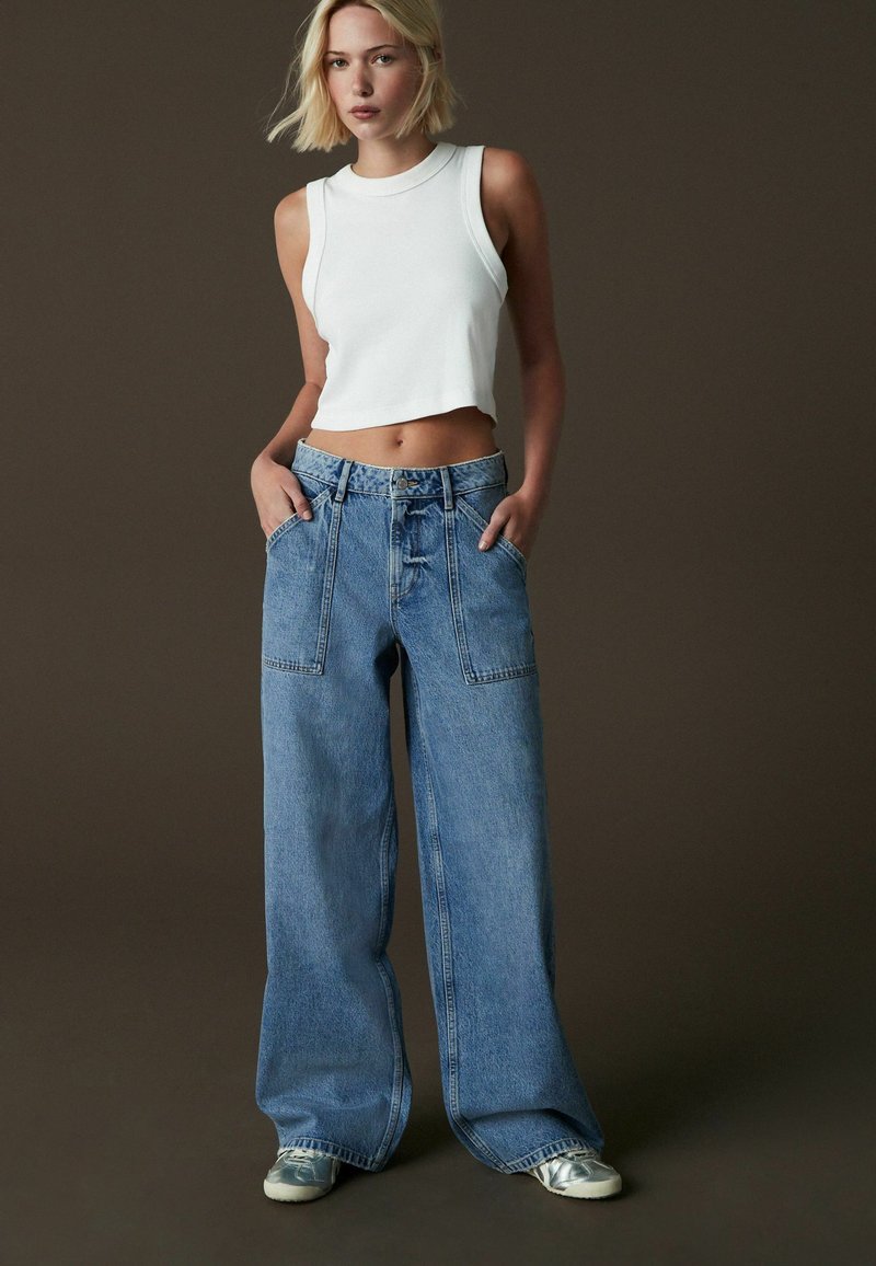 Valkoinen ribbed crop -top, joka on yhdistetty vaaleansinisiin leveisiin farkkuihin, joissa on suuret läppätaskut ja rento istuvuus. Hopeiset tennarit.