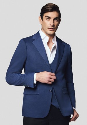 Granatowa marynarka o dopasowanym kroju z teksturowanej tkaniny, z klapami w kształcie notcha i dwoma przednimi kieszeniami, noszona na białej koszuli.