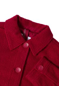 Veste en molleton rouge avec un col, présentant un tissu texturé, deux boutons et une poche avant. L'intérieur possède une doublure rouge contrastante.
