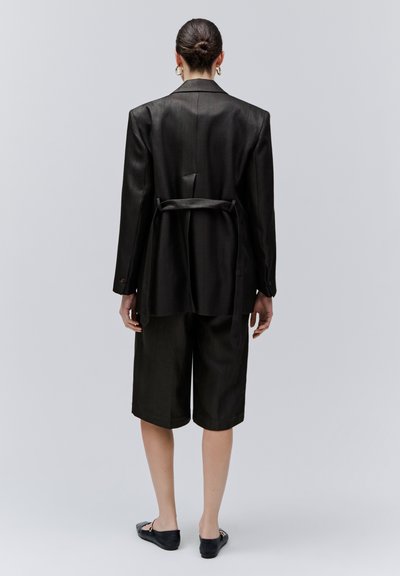 Femme aux cheveux attachés en chignon, portant un blazer noir ceinturé, un short noir longueur genou, des boucles d'oreilles créoles dorées et des chaussures plates noires, debout devant un fond gris.