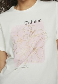 Camiseta de algodón blanca con un gráfico floral rosa con arte en líneas doradas y el texto "S’aimer" sobre una frase en francés más pequeña.