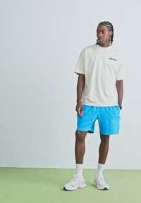 Tricou oversized bej deschis cu logo, asociat cu pantaloni scurți albaștri strălucitori. Sosete atletice albe și adidași completează ținuta.