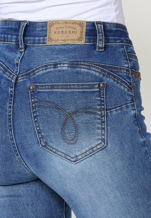 Genska v srednje modrem denimu, z usnjenim oznako, značilno zasnovo šivanja zadnjih žepov in ozkim krojem. Vidna tekstura in podrobnosti.