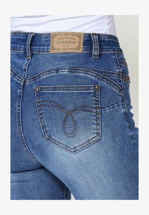 Mėlynų džinsų iš vidutinio mėlyno džinsinio audinio, su odiniu etikete, išskirtiniu užpakalinių kišenių siuvinėjimu ir siaura siluetu. Matoma tekstūra ir detalės.