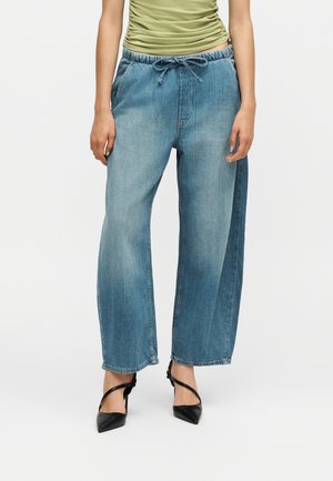 Persoană purtând pantaloni largi din denim albastru cu talie reglabilă cu șnur, top verde-olive drapat şi pantofi negri cu toc ascuțit, stând în picioare.