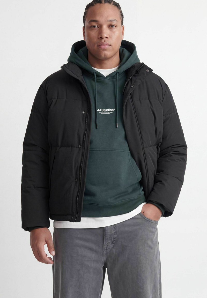 Jeune homme aux cheveux tressés portant une doudoune noire par-dessus un sweat à capuche vert et un pantalon gris, debout avec une main dans la poche.