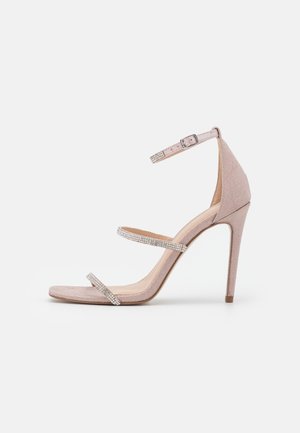 Różowe zamszowe sandały na wysokim obcasie z paskiem w kostce oraz dwoma paskami ozdobionymi cyrkoniami na przodzie stopy. Kwadratowy kształt palców.