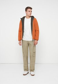 Jachetă portocalie cu fermoar, cu interior verde, tricou alb, pantaloni cargo bej cu multiple buzunare și adidași albi pe un fundal simplu.