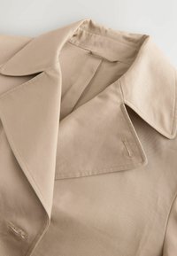 Prim-plan cu gulerul unei trenciuri bej, cu reveruri late și găuri de nasturi vizibile pe material neted.