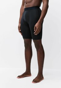 Male model standing barefoot wearing black fitted athletic shorts, shown from mid-torso to feet against a white background.