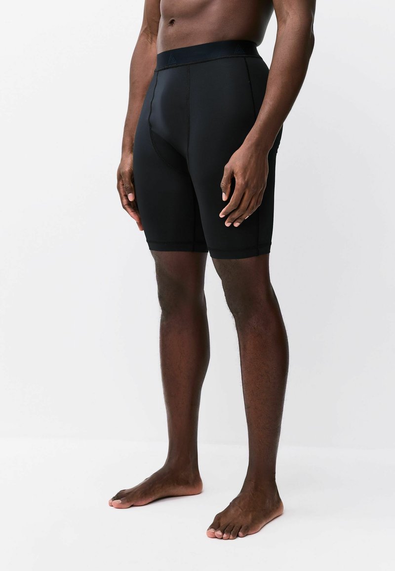 Male model standing barefoot wearing black fitted athletic shorts, shown from mid-torso to feet against a white background.