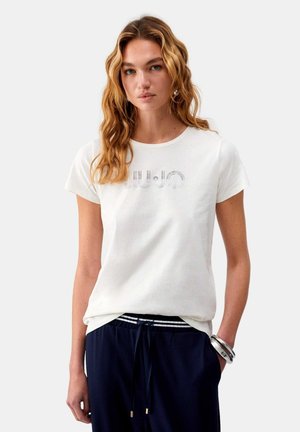 Donna con capelli biondi mossi che indossa una maglietta bianca a maniche corte con un logo decorato in modo sottile, pantaloni blu scuro e braccialetti d'argento al polso sinistro.