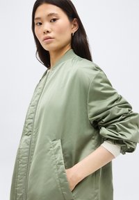 Fiatal nő hosszú, sötét hajjal, világoszöld bomber dzsekit visel, melynek ujjai fodrosak, kezét zsebében tartja, egyszínű háttér előtt.