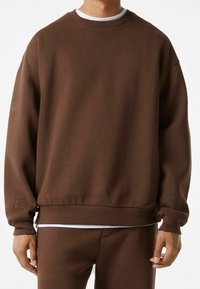 Sweatshirt marron avec un col rond classique, des poignets et un ourlet côtelés. Présente un petit logo sur la partie inférieure de la manche. Tissu doux et texturé.