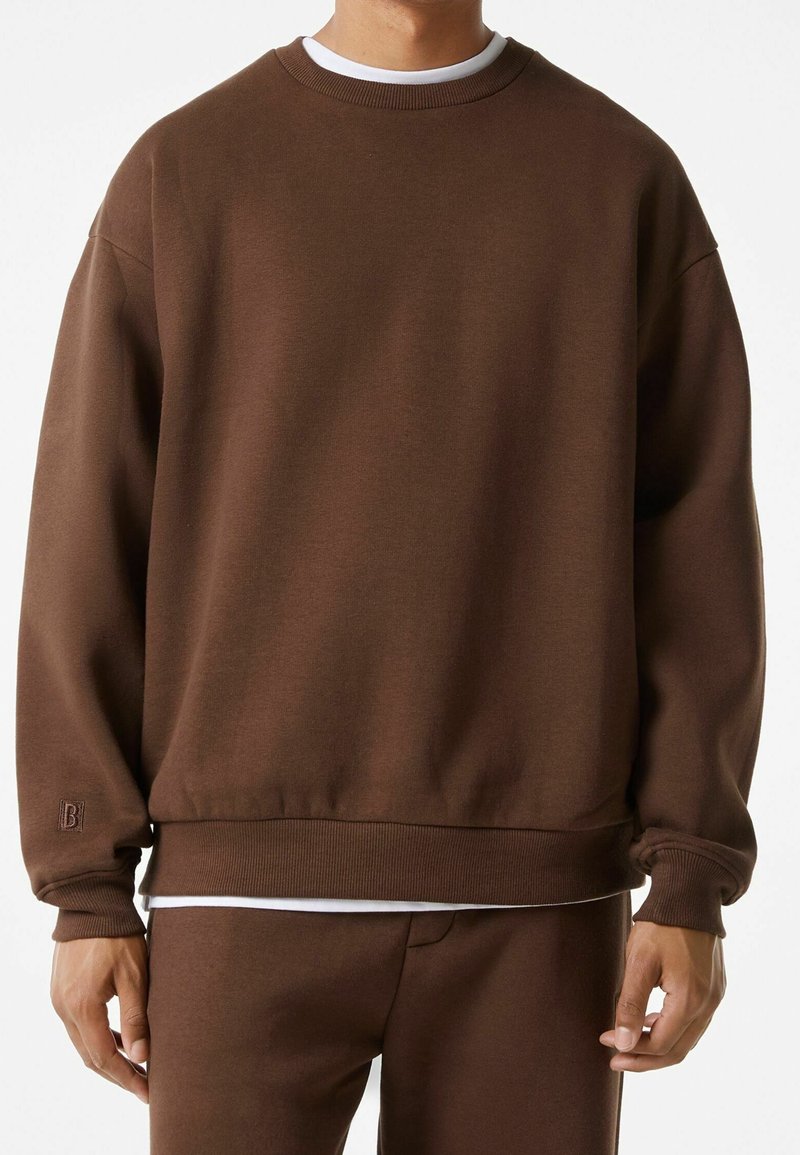 Sweatshirt marron avec un col rond classique, des poignets et un ourlet côtelés. Présente un petit logo sur la partie inférieure de la manche. Tissu doux et texturé.