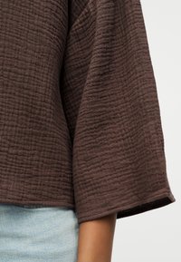 Κοντινό σε χέρι ατόμου που φορεί υφασμάτινο καφέ μακρυμάνικο κορμάκι και ανοιχτό μπλε τζιν παντελόνι μπροστά σε απλό φόντο.