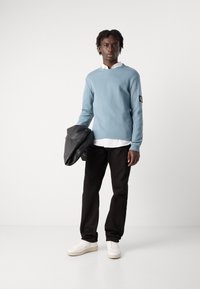 Lys blå teksturert genser, hvit skjorte under, svarte bukser, holder en svart veske, avsluttet med hvite sneakers. Enkelt design og sporty utseende.