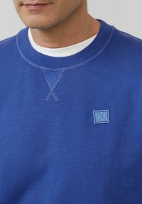 Sweatshirt bleu à col rond et col côtelé. Présente un petit patch logo brodé et des détails de couture contrastants. Texture douce.