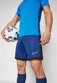 Sinine Nike lühikeste varrukatega särk mustade külgpaneelidega, millele on sobitatud tumesinised lühikesed püksid, millel on oranž logo. Valge jalgpall siniste aktsentidega.