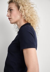 Marineblå polo skjorte med korte ermer og krave, laget av glatt materiale. Har en subtil logo på brystet. Krøllete hår synlig.