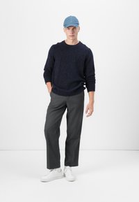 Granatowy sweter o melanżowym splocie, szare spodnie o szerokich nogawkach i białe sneakersy. Całość dopełnia casualowa czapka dżinsowa. Proste białe tło.