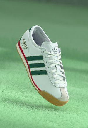 White Adidas sneaker with green stripes, red accent, gum sole, and "Italy" text, floating above green grass background.