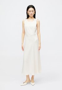 Μακρύ, χωρίς μανίκια, κρεμ φόρεμα σε λεία ύφανση. Διαθέτει τετράγωνη λαιμόκοψη και πέφτει χαλαρά μέχρι το μισό της κνήμης, συνδυασμένο με λευκά μυτερά μπαλαρίνες.